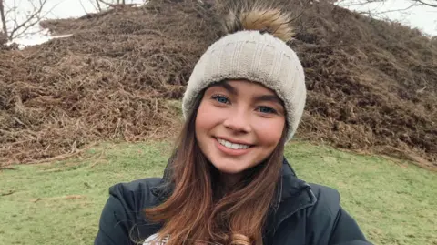 Family handout A selfie of Jaysley Beck, who has long brown hair and is wearing a black jacket and a cream-coloured beanie hat. She is pictured standing on a grassy hill on a cold winter's day.
