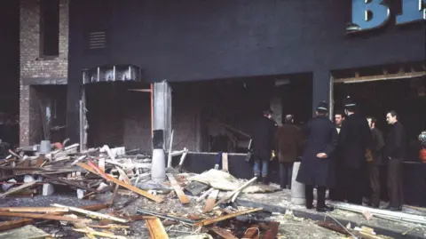 BBC Wreckage including splintered wood and rubble on the pavement outside a building whose front has been blown out. Two policemen in uniform and helmets stand with their backs to camera in a group with three other men in dark clothing. Two men in coats stand looking inside the ruined building with their backs to the camera.
