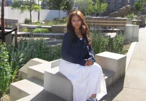 Alisha Miah A woman in a navy cardigan and long white skirt sits outside on a modern-looking concrete bench in front of some plants. She is smiling at the camera