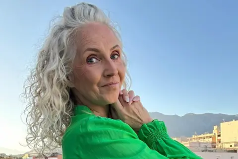 Rhian Davies Rhian has curly light blonde/grey hair. In this photographs she is smiling at the camera and wearing an emerald green top. She is in a sunny environment. 