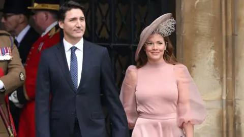 Getty Images Justin Trudeau and Sophie Grégoire Trudeau arrive at Westminster Abbey for the Coronation of King Charles III and Queen Camilla on May 06, 2023 in London, England. Trudeau is wearing a navy blue suit with a polka-dotted navy blue tie, while Sophie is wearing a blush pink dress with a high neck and chiffon, billowing sleeves. She is wearing a matching fascinator on her head. The two are holding hands and smiling. 
