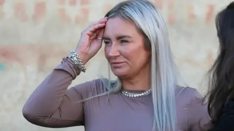 Reuters Lyanne Kennedy speaking to reporters outside Tbilisi City Court. She has long, dyed platinum-blonde hair with dark roots showing and is wearing silver necklaces, bracelets and rings.  She has on a mauve, long-sleeved t-shirt. Graffiti is on the wall behind her.